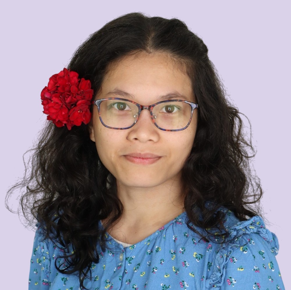 Portrait of Sythong Run. She wear blue flowert buttonup and blue glasses. She wears her wavy hair down with a red flower pinned to her left ear.